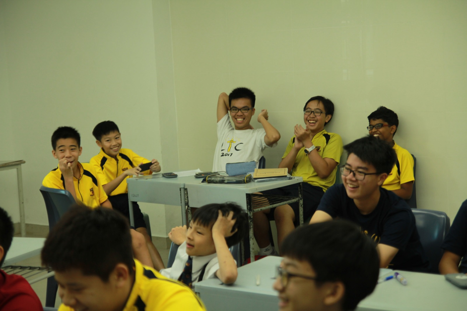 A group of students in yellow tops sit around a table in a classroom, smiling and reacting enthusiastically, some raising their hands, while looking forward..